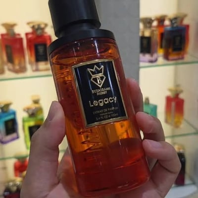 Bujairami Legacy by Bujairami 100ml Extrait — An amber-colored glass perfume bottle with a black cap and a black label featuring gold text and a gold crest. | Bujairami Legacy by Bujairami 100ml Extrait — Botol kaca berwarna ambar dengan penutup hitam dan label hitam yang menampilkan teks emas dan lambang emas.