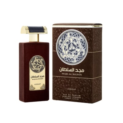 Asdaaf Majd Al Sultan — A dark brown velvet-textured perfume bottle with a gold and black cap stands next to its matching brown box, both featuring a circular Celtic knot design. | Asdaaf Majd Al Sultan — Botol minyak wangi bertekstur baldu coklat gelap dengan penutup emas dan hitam berdiri di sebelah kotak coklat
