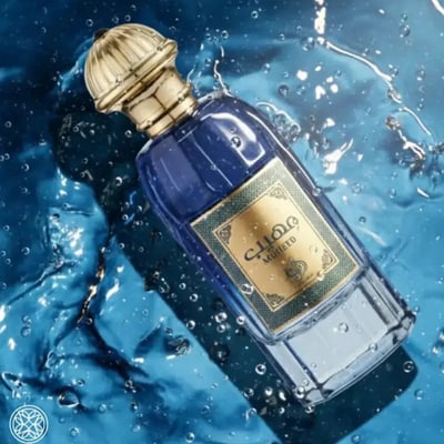 Al Wataniah Muheeb Perfume Man by Al Wataniah 100ml — A dark blue perfume bottle with a gold cap sits next to its matching dark blue box with ornate gold Arabic calligraphy and floral designs. | Al Wataniah Muheeb Perfume Man by Al Wataniah 100ml — Botol minyak wangi biru gelap dengan penutup emas terletak di sebelah