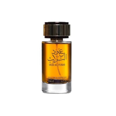Ard Al Zaafaran Oud Al Turas — A clear glass perfume bottle with golden cap and a dark dried plant inside, next to a gold and maroon ornate box. | Botol minyak wangi kaca jernih dengan penutup emas dan tumbuhan kering gelap di dalamnya, bersebelahan dengan kotak berhias emas dan merah marun.
