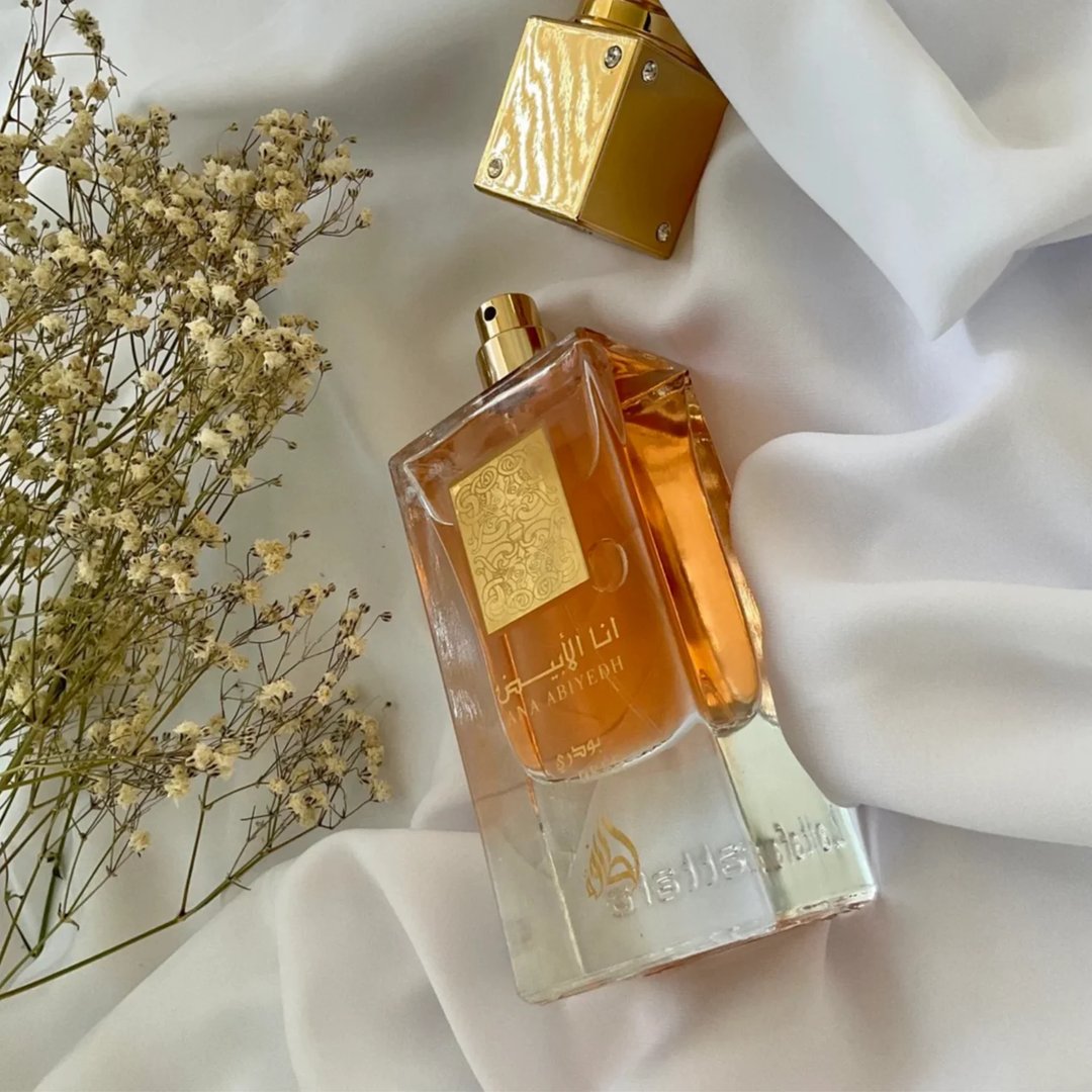 Lattafa Ana Abiyedh Poudree by Lattafa 60ml — A white perfume box with gold accents and Arabic calligraphy stands next to a clear glass perfume bottle with a gold cap and intricate gold detailing on the bottle. | Lattafa Ana Abiyedh Poudree by Lattafa 60ml — Kotak minyak wangi putih dengan aksen emas dan kaligrafi