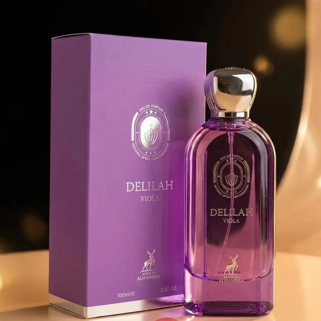 Maison Alhambra Delilah Viola 100ml — A purple glass perfume bottle with a gold cap stands next to its matching purple box, both featuring gold embossed details and a deer logo. | Maison Alhambra Delilah Viola 100ml — Botol kaca ungu berdiri di sebelah kotak ungu sepadan, kedua-duanya menampilkan butiran emas timbul
