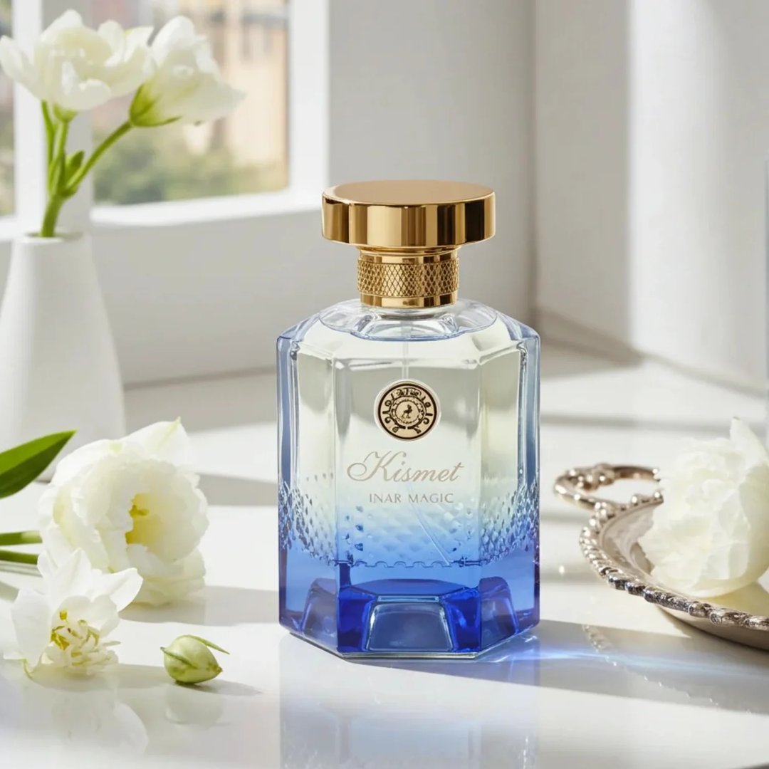 Maison Alhambra Kismet Lunar Magic 100ml — A clear glass perfume bottle with a gold cap and blue gradient base sits next to its matching blue and gold box on a white background. | Maison Alhambra Kismet Lunar Magic 100ml — Botol minyak wangi kaca jernih dengan penutup emas dan tapak gradien biru terletak di sebelah