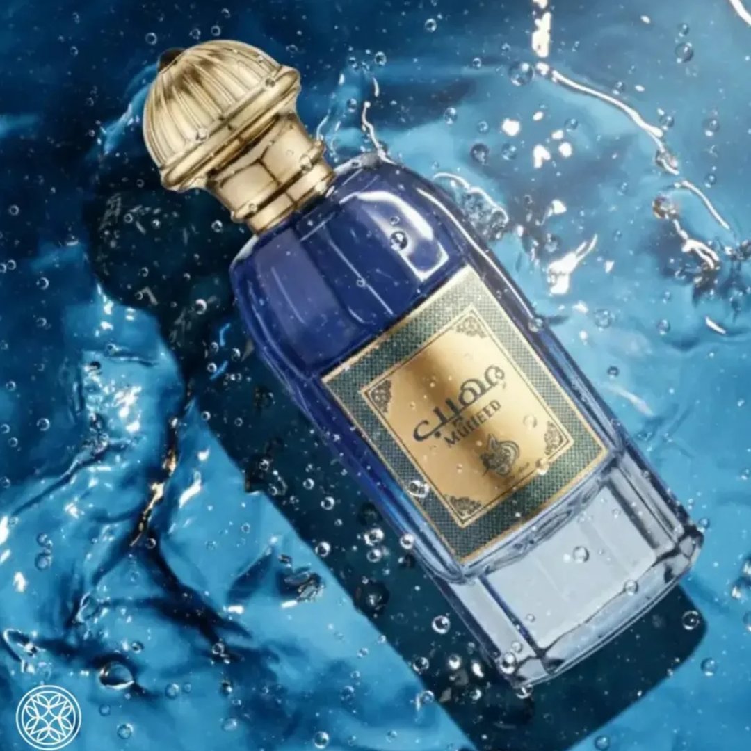 Al Wataniah Muheeb Perfume Man by Al Wataniah 100ml — A dark blue perfume bottle with a gold cap sits next to its matching dark blue box with ornate gold Arabic calligraphy and floral designs. | Al Wataniah Muheeb Perfume Man by Al Wataniah 100ml — Botol minyak wangi biru gelap dengan penutup emas terletak di sebelah