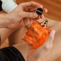 Rayhaan Adonis Icarus Perfume — A gold embossed box sits next to a square amber glass perfume bottle with a gold cap, both featuring intricate embossed patterns. | Rayhaan Adonis Icarus Perfume — Kotak emas bertekstur duduk bersebelahan botol minyak wangi kaca berwarna ambar berbentuk segi empat dengan penutup emas