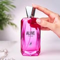 Maison Alhambra Alive Now — A bright pink faceted glass perfume bottle with a silver geometric cap stands next to its matching pink box, which features a dotted pattern and the brand logo. | Maison Alhambra Alive Now — Botol kaca berwarna merah jambu terang berbentuk faset dengan penutup geometri perak berdiri di