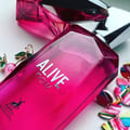 Maison Alhambra Alive Now — A bright pink faceted glass perfume bottle with a silver geometric cap stands next to its matching pink box, which features a dotted pattern and the brand logo. | Maison Alhambra Alive Now — Botol kaca berwarna merah jambu terang berbentuk faset dengan penutup geometri perak berdiri di