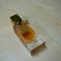 Lattafa Ana Abiyedh Poudree by Lattafa 60ml — A white perfume box with gold accents and Arabic calligraphy stands next to a clear glass perfume bottle with a gold cap and intricate gold detailing on the bottle. | Lattafa Ana Abiyedh Poudree by Lattafa 60ml — Kotak minyak wangi putih dengan aksen emas dan kaligrafi