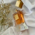 Lattafa Ana Abiyedh Poudree by Lattafa 60ml — A white perfume box with gold accents and Arabic calligraphy stands next to a clear glass perfume bottle with a gold cap and intricate gold detailing on the bottle. | Lattafa Ana Abiyedh Poudree by Lattafa 60ml — Kotak minyak wangi putih dengan aksen emas dan kaligrafi