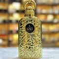 Lattafa Bayaan Eau De Parfum — A gold-colored perfume bottle with ornate Arabic script and a gold ring handle sits next to its matching gold box, also adorned with script and a circular logo. | Lattafa Bayaan Eau De Parfum — Botol minyak wangi berwarna emas dengan skrip Arab berhias dan pemegang cincin emas duduk
