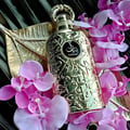 Lattafa Bayaan Eau De Parfum — A gold-colored perfume bottle with ornate Arabic script and a gold ring handle sits next to its matching gold box, also adorned with script and a circular logo. | Lattafa Bayaan Eau De Parfum — Botol minyak wangi berwarna emas dengan skrip Arab berhias dan pemegang cincin emas duduk