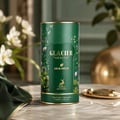 Maison Alhambra Glacier Heaven 100ml — Green floral patterned cylindrical box and a clear glass perfume bottle with green liquid and gold cap, set against a white background. | Maison Alhambra Glacier Heaven 100ml — Kotak silinder hijau bermotif bunga dan botol minyak wangi kaca jernih dengan cecair hijau dan penutup