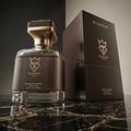 Bujairami Kingsman — A clear glass perfume bottle with a black leather band and gold accents sits next to its dark brown