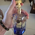 Maison Alhambra Kismet Lunar Magic 100ml — A clear glass perfume bottle with a gold cap and blue gradient base sits next to its matching blue and gold box on a white background. | Maison Alhambra Kismet Lunar Magic 100ml — Botol minyak wangi kaca jernih dengan penutup emas dan tapak gradien biru terletak di sebelah