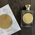 Ard Al Zaafaran Mithqal 100ml — A clear glass perfume bottle with gold liquid and a circular gold emblem, next to its white and gold patterned box with a matching emblem. | Ard Al Zaafaran Mithqal 100ml — Botol minyak wangi kaca jernih dengan cecair emas dan lambang emas bulat, bersebelahan dengan kotak corak putih