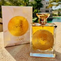 Ard Al Zaafaran Mithqal 100ml — A clear glass perfume bottle with gold liquid and a circular gold emblem, next to its white and gold patterned box with a matching emblem. | Ard Al Zaafaran Mithqal 100ml — Botol minyak wangi kaca jernih dengan cecair emas dan lambang emas bulat, bersebelahan dengan kotak corak putih