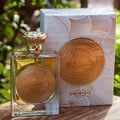 Ard Al Zaafaran Mithqal 100ml — A clear glass perfume bottle with gold liquid and a circular gold emblem, next to its white and gold patterned box with a matching emblem. | Ard Al Zaafaran Mithqal 100ml — Botol minyak wangi kaca jernih dengan cecair emas dan lambang emas bulat, bersebelahan dengan kotak corak putih