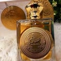 Ard Al Zaafaran Mithqal 100ml — A clear glass perfume bottle with gold liquid and a circular gold emblem, next to its white and gold patterned box with a matching emblem. | Ard Al Zaafaran Mithqal 100ml — Botol minyak wangi kaca jernih dengan cecair emas dan lambang emas bulat, bersebelahan dengan kotak corak putih