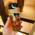 Lattafa Musamam Eau De Parfum 100ml by Lattafa — A gold snake wraps around a clear glass perfume bottle filled with amber liquid, next to a textured brown cylindrical box. | Lattafa Musamam Eau De Parfum 100ml by Lattafa — Ular emas melilit botol minyak wangi kaca jernih berisi cecair ambar, bersebelahan kotak
