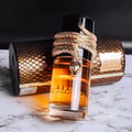 Lattafa Musamam Eau De Parfum 100ml by Lattafa — A gold snake wraps around a clear glass perfume bottle filled with amber liquid, next to a textured brown cylindrical box. | Lattafa Musamam Eau De Parfum 100ml by Lattafa — Ular emas melilit botol minyak wangi kaca jernih berisi cecair ambar, bersebelahan kotak