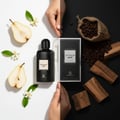 Optimum Noir centered in a split-tone scene: bright pear/orange blossom vs dark coffee/cedar; Malaysian model hands; clear product.