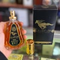Ard Al Zaafaran Qaed Al Fursan — A gold-capped glass perfume bottle with amber liquid sits next to its black and gold box featuring a running horse graphic. | Ard Al Zaafaran Qaed Al Fursan — Botol kaca bertutup emas dengan cecair berwarna ambar duduk bersebelahan dengan kotak hitam dan emasnya yang menampilkan grafik