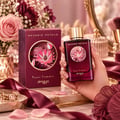 Zimaya Reverie Petals by Zimaya 100ml EDP Women — A woman's hand with dark red nails reaches for the gold cap of a deep red perfume bottle, next to its matching box, surrounded by dark red flowers and delicate white blossoms on a marble surface.