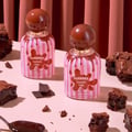Grandeur Tubbees Chocolate Fudge — A perfume bottle with a chocolate-colored spherical cap and pink and white striped body, with chocolate drips cascading down. Next to it is the product box with the same design. | Grandeur Tubbees Chocolate Fudge — Botol minyak wangi dengan penutup bulat berwarna coklat dan badan